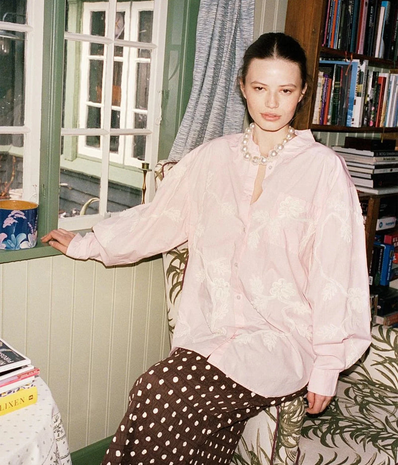 A woman seated indoors wearing a light pink embroidered blouse and dark brown polka-dot trousers, with a pearl necklace, in front of a window and bookshelves.