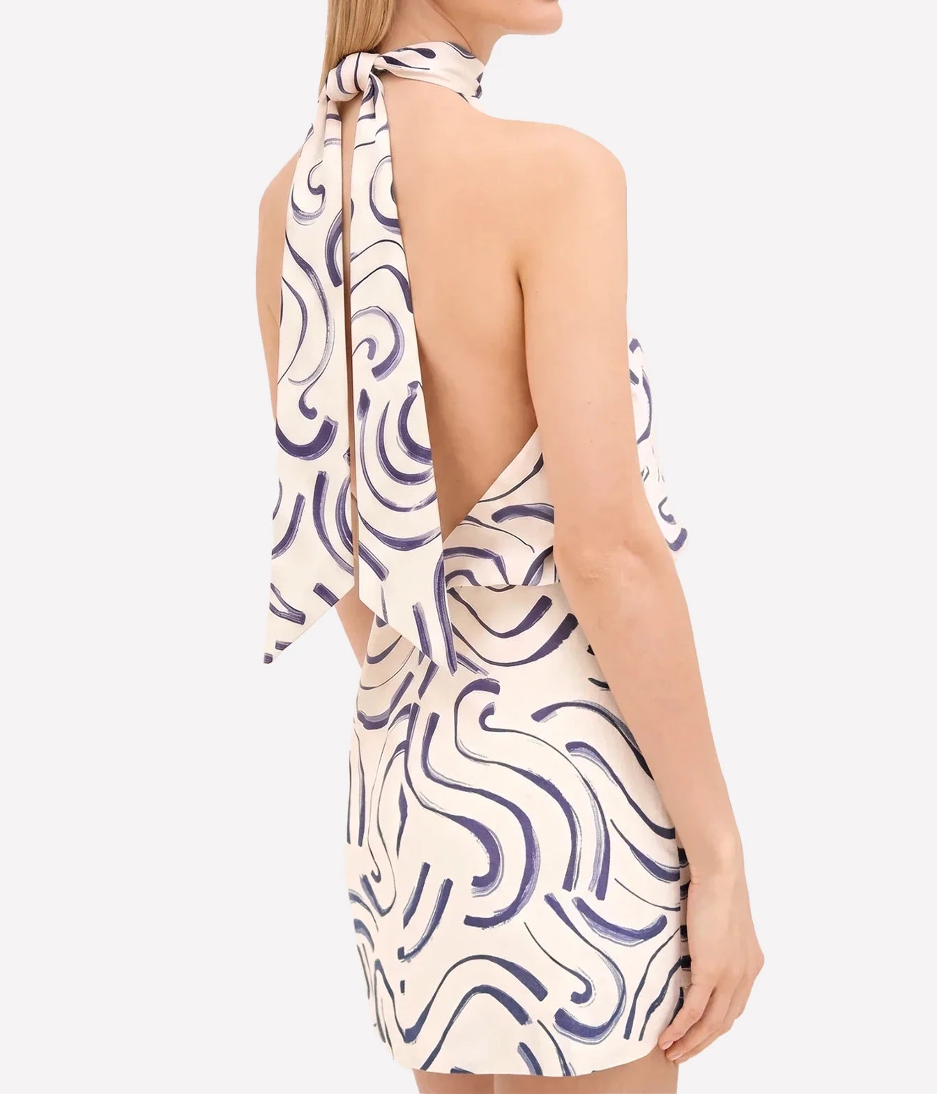 A woman shown from the back wearing a halter-neck mini dress with a cream base and a dark purple abstract wavy pattern. The dress features a tie-shoulder design and a wrap-style back cutout.