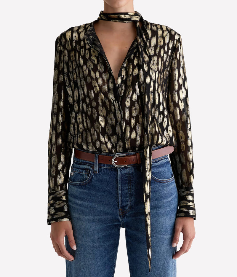 Model wearing the AG Lora Tie Neck Shirt in Leopard Lounge tucked into mid-blue jeans with a brown leather belt, the tie neck knotted loosely with long ends hanging down the front