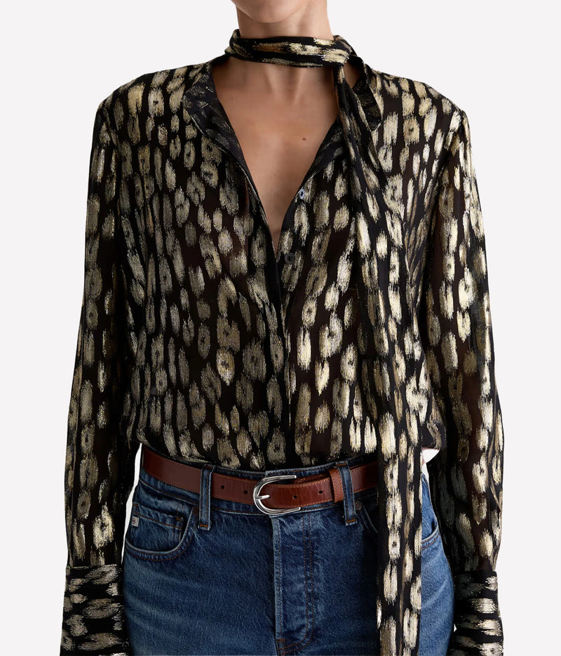 Close front view of a model wearing the AG Lora Tie Neck Shirt in Leopard Lounge with the tie wrapped at the neck and an open V-neckline, tucked into blue jeans with a brown leather belt