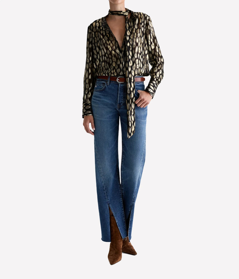 Full-length view of a model wearing the AG Lora Tie Neck Shirt in Leopard Lounge tucked into blue wide-leg jeans with brown boots, the tie draped loosely around the neck