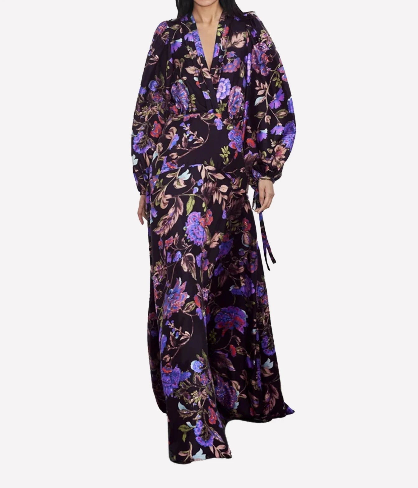 A woman wearing a long-sleeved, floor-length floral dress with a dark base and purple, pink, and green botanical print, shown against a plain light gray background.