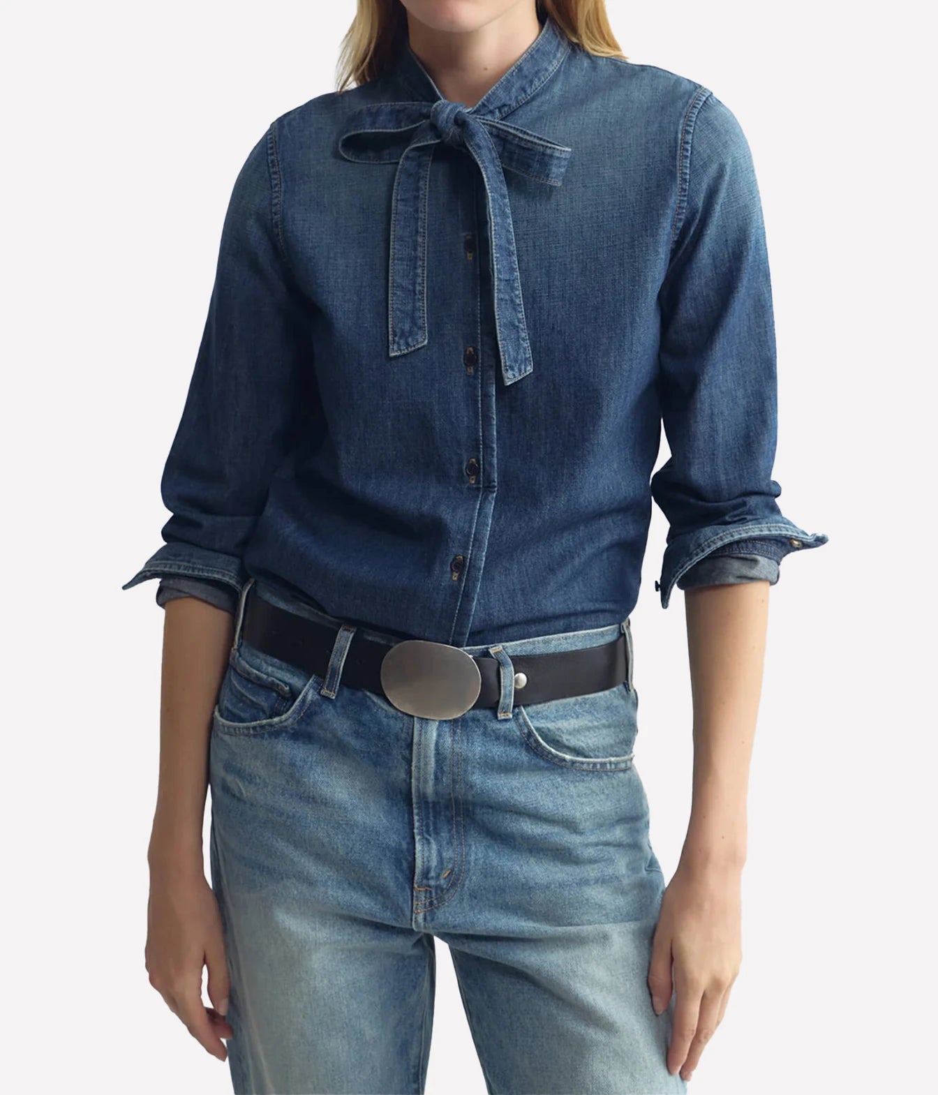 A model wearing a dark blue denim shirt with a tie-neck detail, paired with light-wash blue jeans and a black belt with a silver oval buckle, against a plain light gray background.