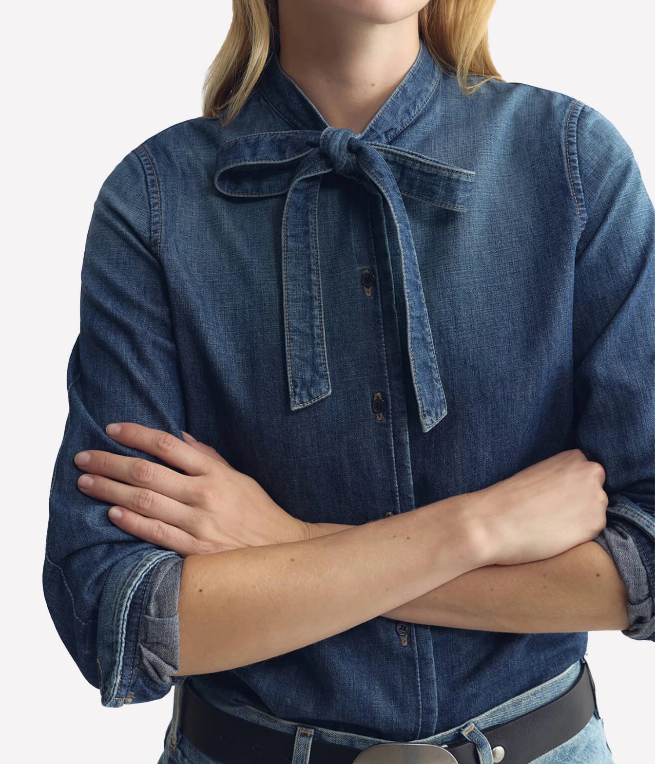 A close-up of a model wearing a blue denim button-up shirt with a tie-neck detail and rolled sleeves, paired with a black belt against a light gray background.