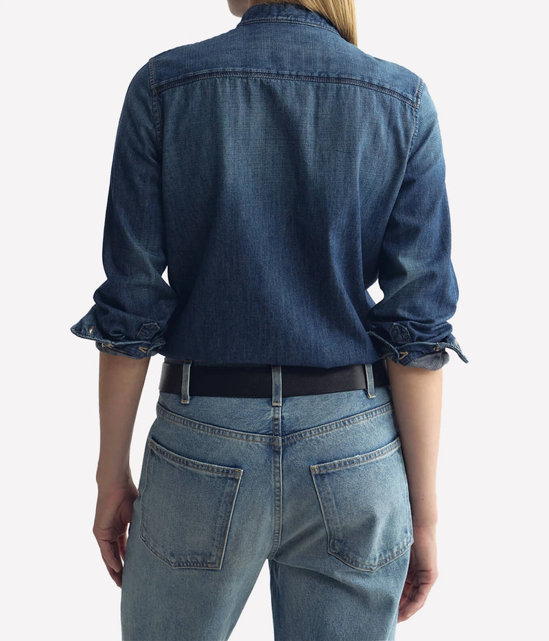 Back view of a person wearing a dark blue denim button-up shirt with rolled sleeves and light blue jeans, against a plain light gray background.
