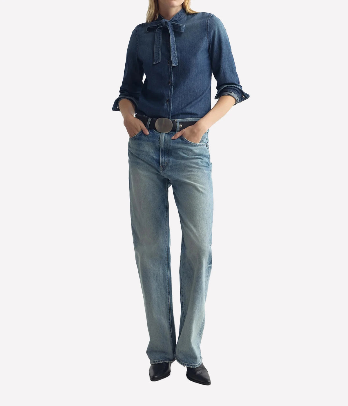 A model wearing a dark blue denim long-sleeve shirt with a tie collar and light-wash blue denim jeans, styled with a silver belt and black shoes, against a plain white background.