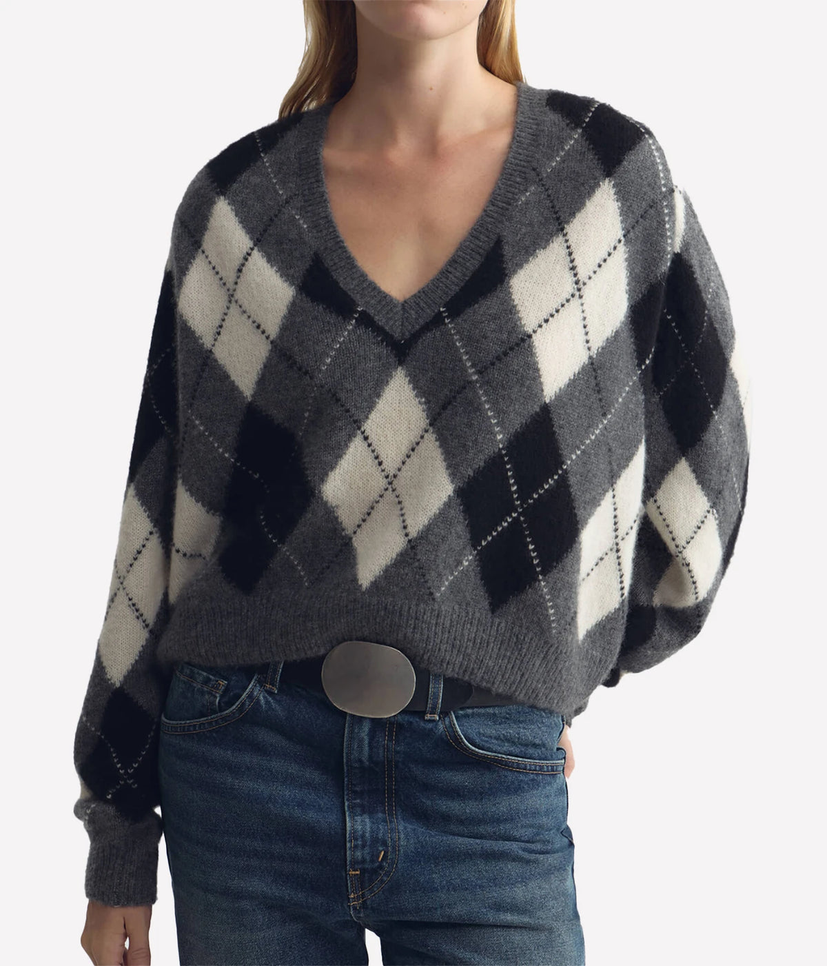 A woman wearing a gray, black, and off-white argyle knit sweater with blue jeans and a silver belt, shown from the neck down against a light gray background.
