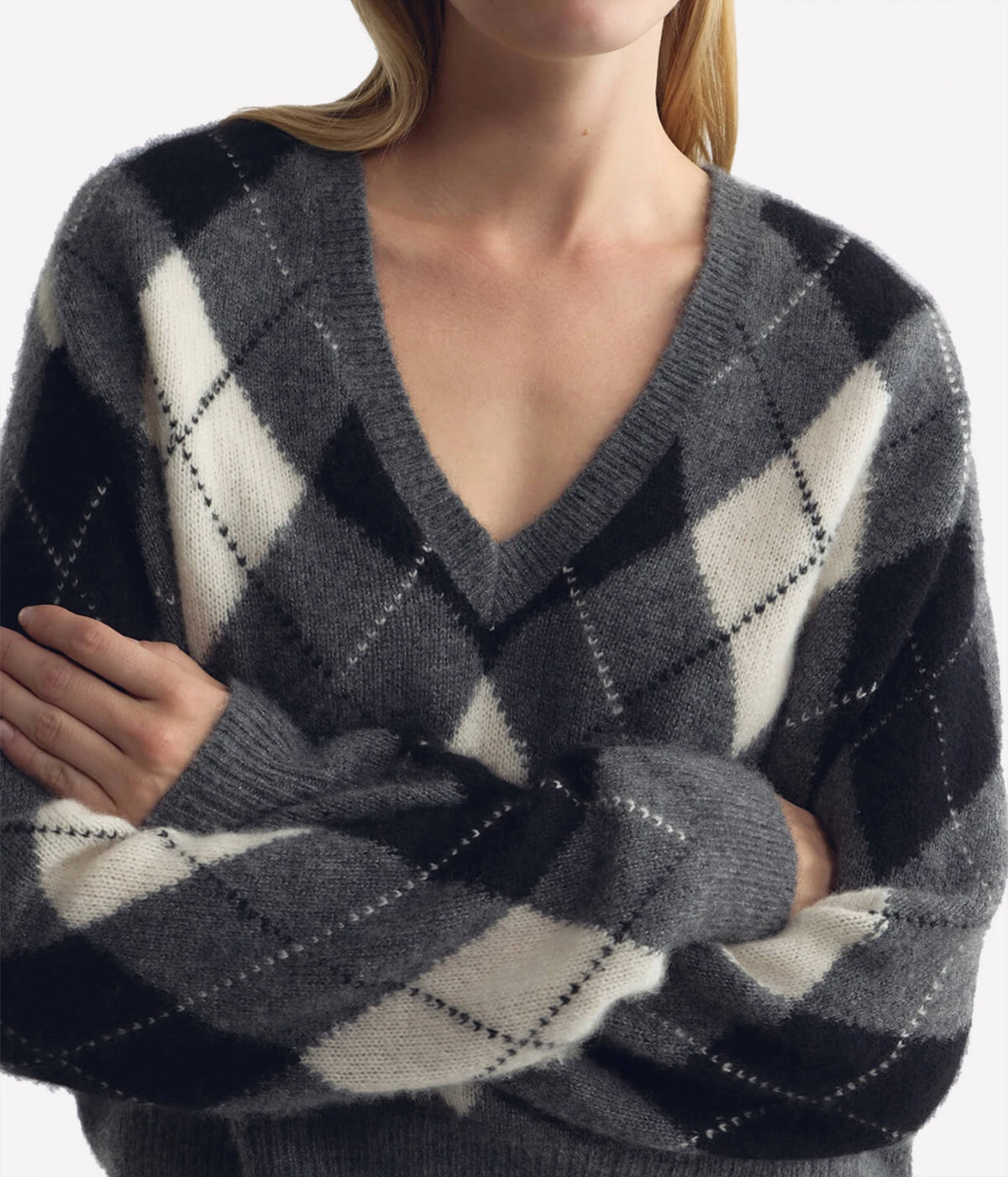 A close-up of a woman wearing a gray, black, and off-white argyle V-neck sweater, with her arms crossed.