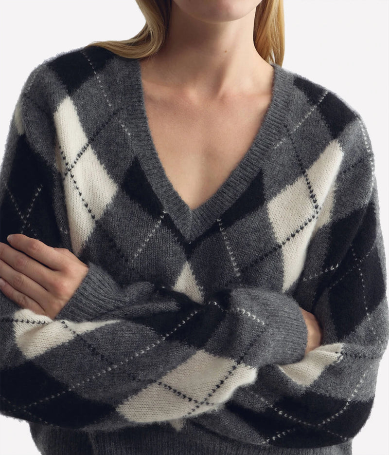 A close-up of a woman wearing a gray, black, and off-white argyle V-neck sweater, with her arms crossed.