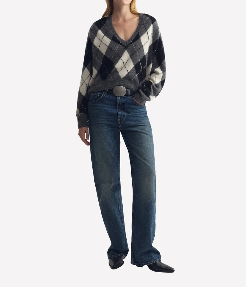 A model wearing a black, gray, and white argyle sweater vest over a dark blue straight-leg denim jacket, styled with a silver belt and black pointed shoes, against a plain white background.