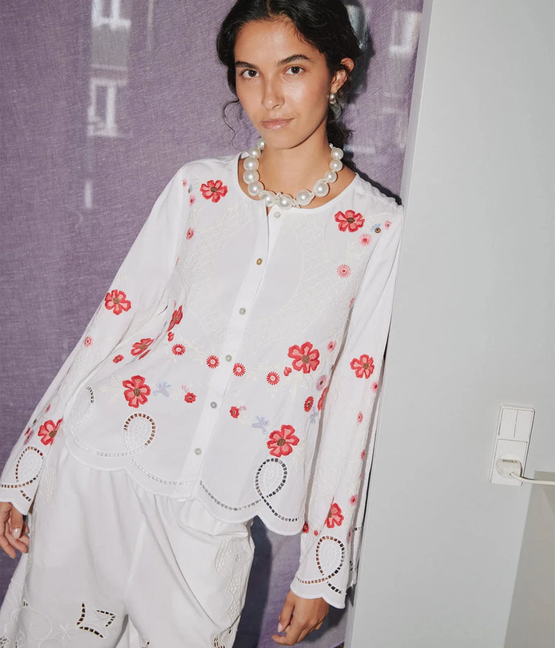 A woman wearing a white long-sleeve blouse with red floral embroidery and scalloped edges, paired with white embroidered pants, standing against a purple textured backdrop.