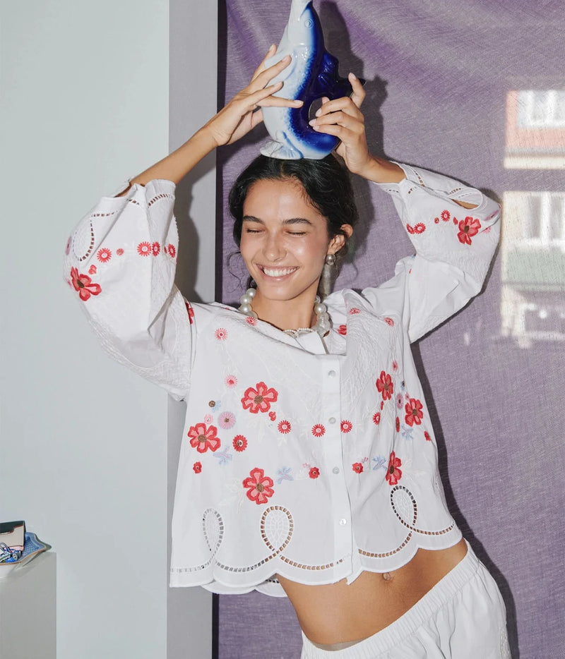 A woman wearing a white long-sleeve blouse with red floral embroidery and scalloped edges, balancing a blue and white shell on her head in an indoor setting.