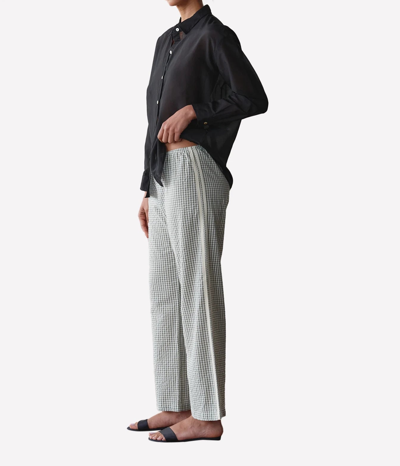 A model shown from the side wearing a black button-down shirt, light gray and white gingham wide-leg trousers, and black slide sandals, set against a plain light gray background.