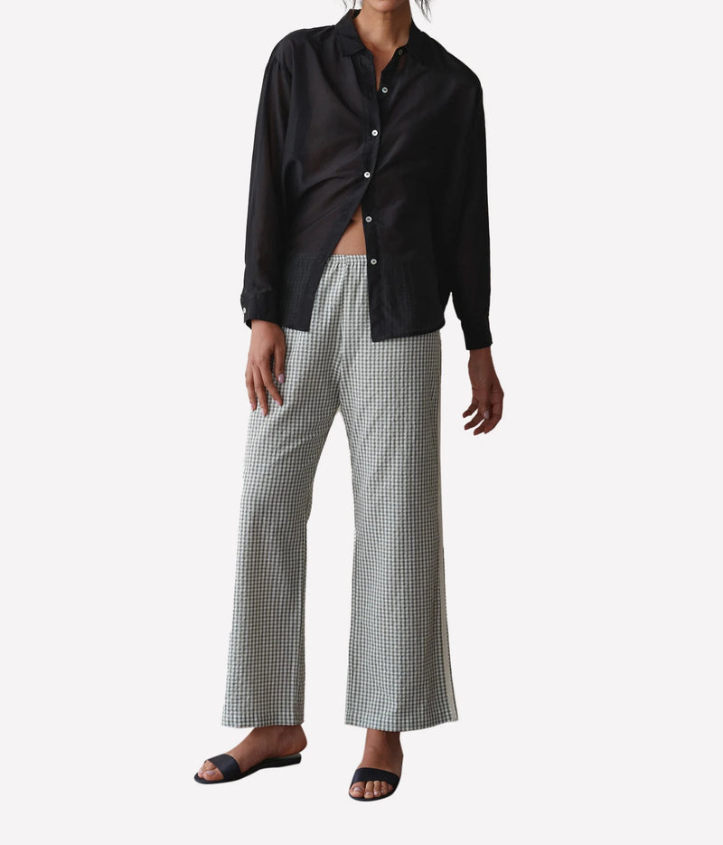 A model wearing a black button-up shirt and wide-leg cropped trousers with a small green and white gingham pattern, paired with black slide sandals, against a light gray studio background.