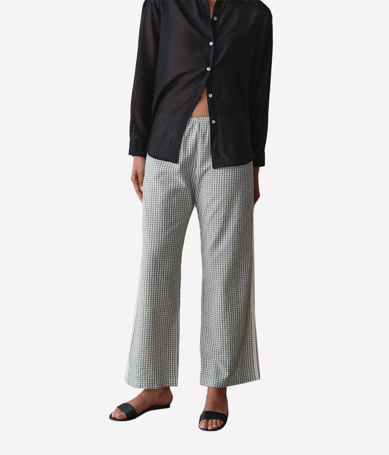 A model wearing a black button-down shirt and light gray and black gingham wide-leg trousers, paired with black slide sandals, against a plain light gray background.