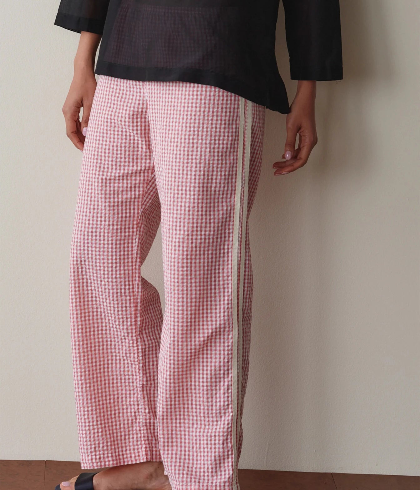 Model wearing pink gingham check pants, side view leaning against a wall to show the straight-leg silhouette and contrast side stripe.