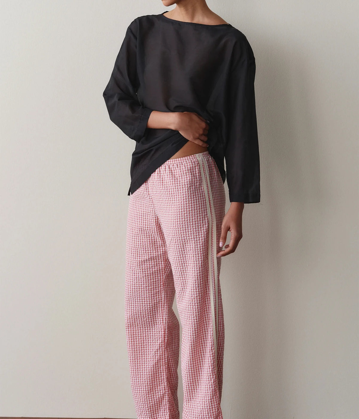 Model wearing pink gingham check pants with a black long-sleeve top, side profile view against a wall.