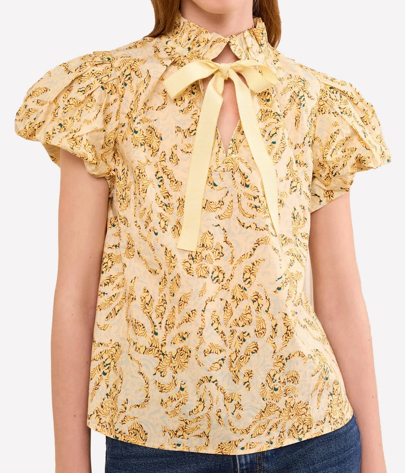 A woman wearing a pale yellow blouse with a gold and green paisley pattern, featuring a keyhole neckline with a yellow ribbon bow, short puff sleeves, and blue jeans.
