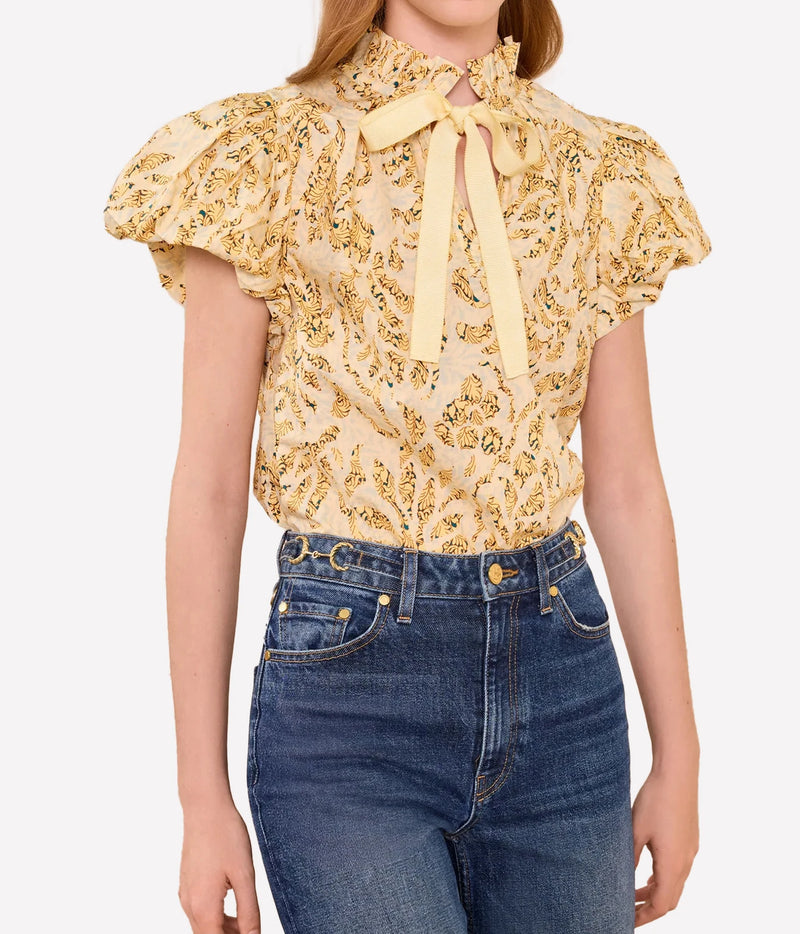 A woman wearing a pale yellow blouse with a yellow and green floral pattern, ruffled short sleeves, and a tie-neck bow, paired with blue jeans against a light gray background.