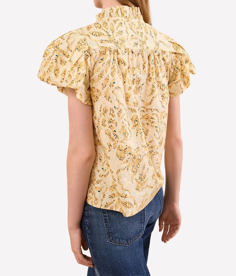 A woman wearing a cream-colored blouse with a yellow and green paisley pattern, featuring ruffled short sleeves and a smocked neck, paired with blue jeans against a light gray background.