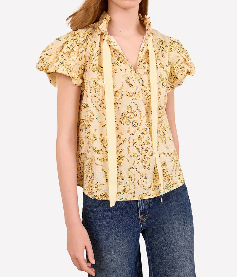 A woman wearing a pale yellow short-sleeve blouse with a yellow and green paisley pattern, ruffled shoulders, and tie details at the collar, paired with blue jeans against a light gray background.