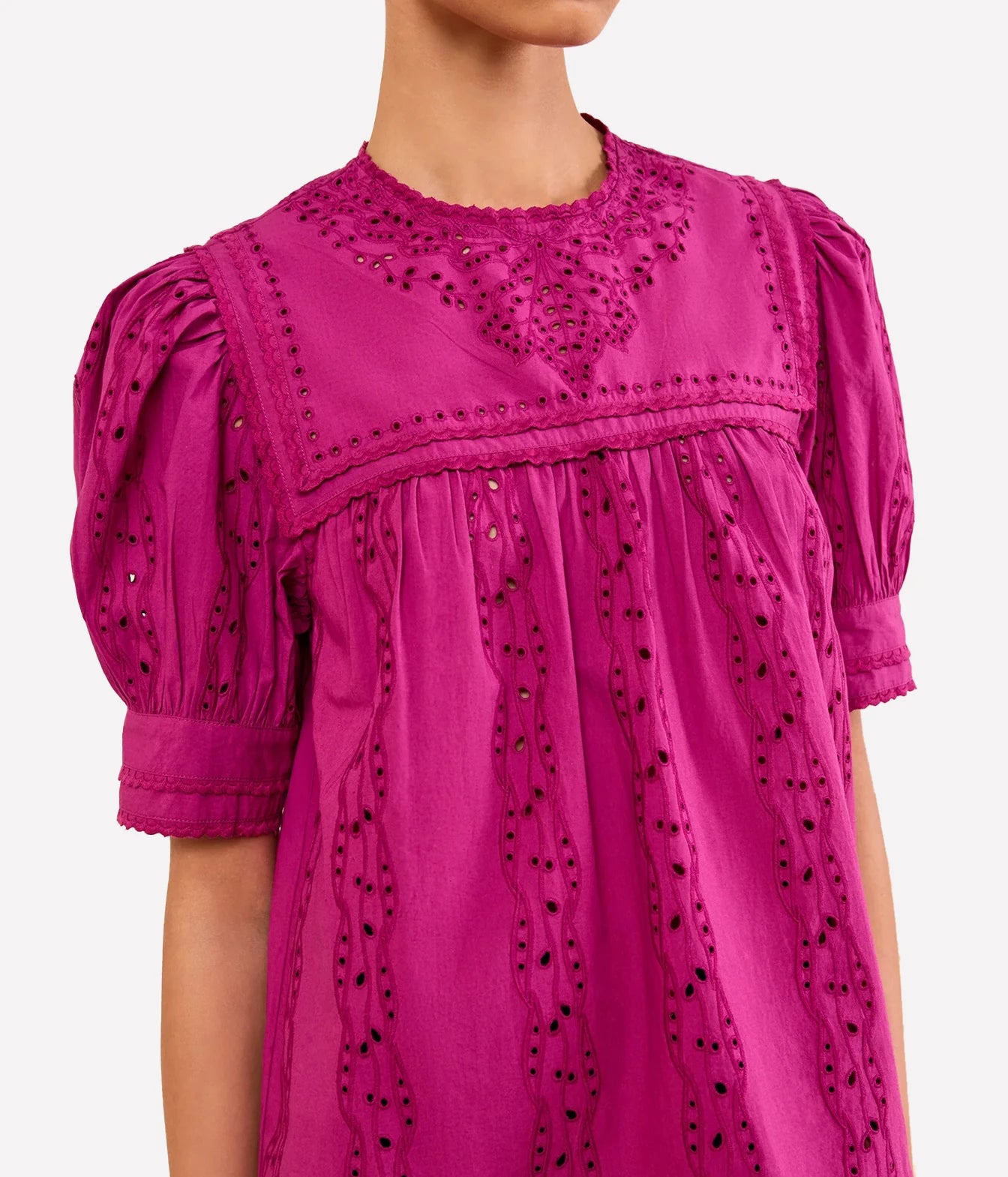 A close-up of a model wearing a bright magenta pink eyelet blouse with short puff sleeves and a gathered neckline, shown against a plain light gray background.
