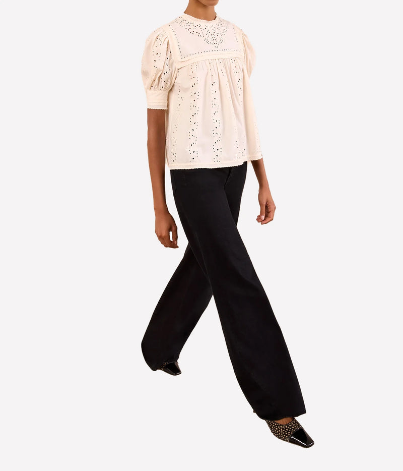 A model wearing a cream-colored eyelet lace blouse with short puff sleeves and black wide-leg trousers, shown from the waist up against a light gray background.