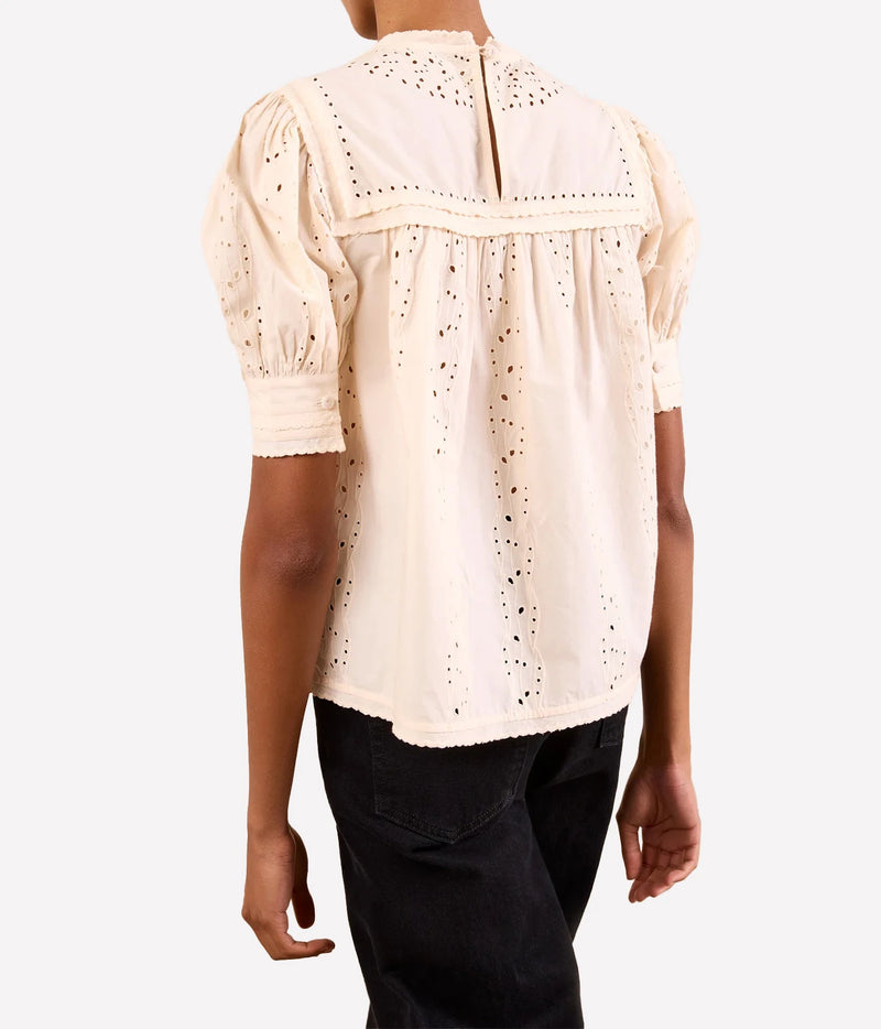 A person shown from the back wearing a cream-colored eyelet blouse with puff sleeves and black pants, photographed against a light gray studio background.