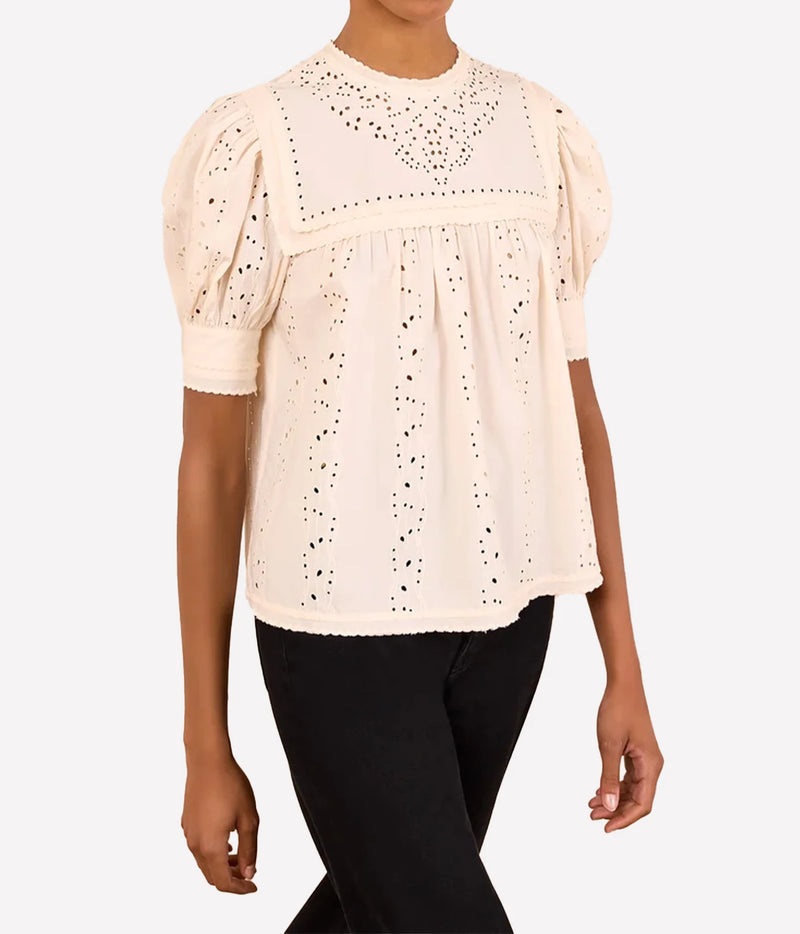 A woman wearing a cream-colored eyelet blouse with puff sleeves and black pants, shown from the neck down against a light gray background.