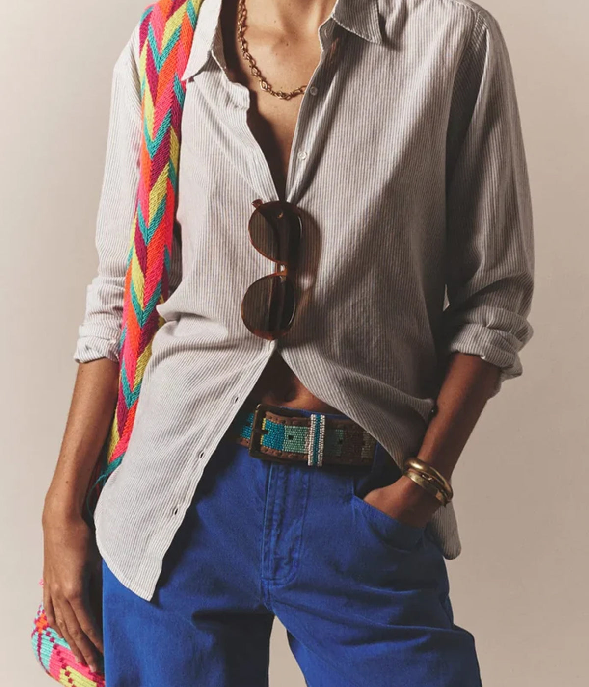 Model wearing the Xirena Beau Shirt in Windward Stripe unbuttoned over cobalt blue jeans with a woven belt, gold chain necklace, gold cuff, sunglasses hooked at the placket and a multicoloured woven crossbody bag