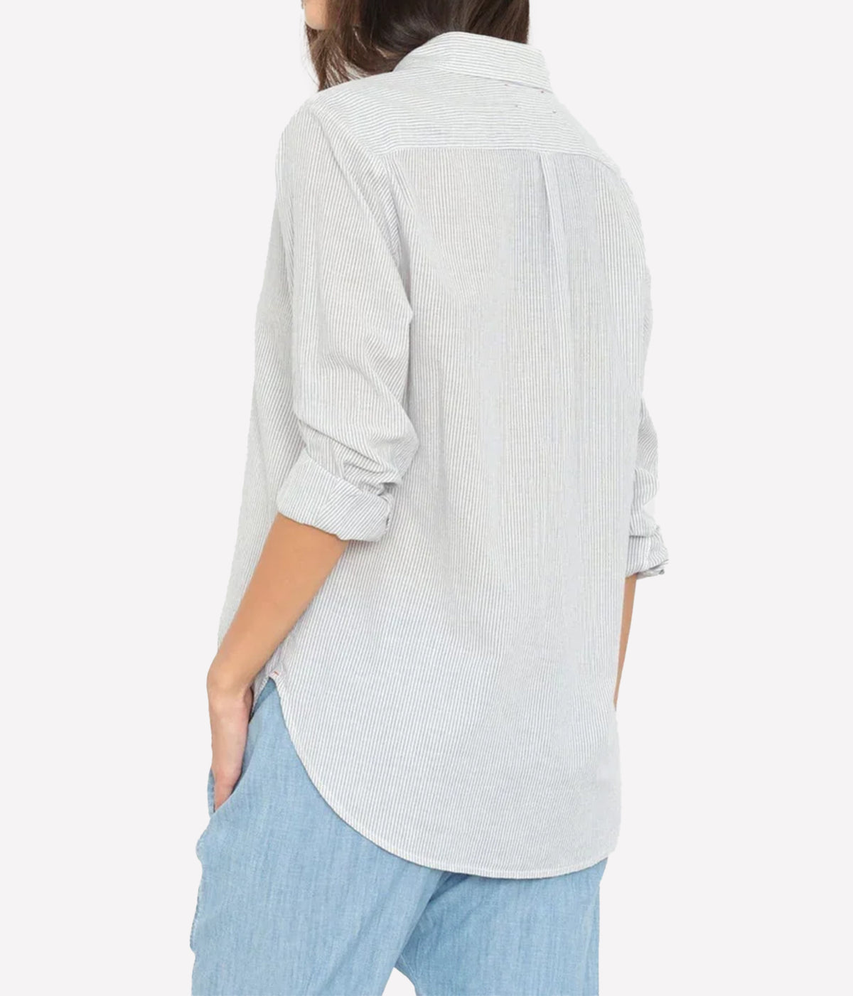 Back view of a model wearing the Xirena Beau Shirt in Windward Stripe untucked over chambray pants, showing the back yoke, rolled sleeves and curved shirttail hem