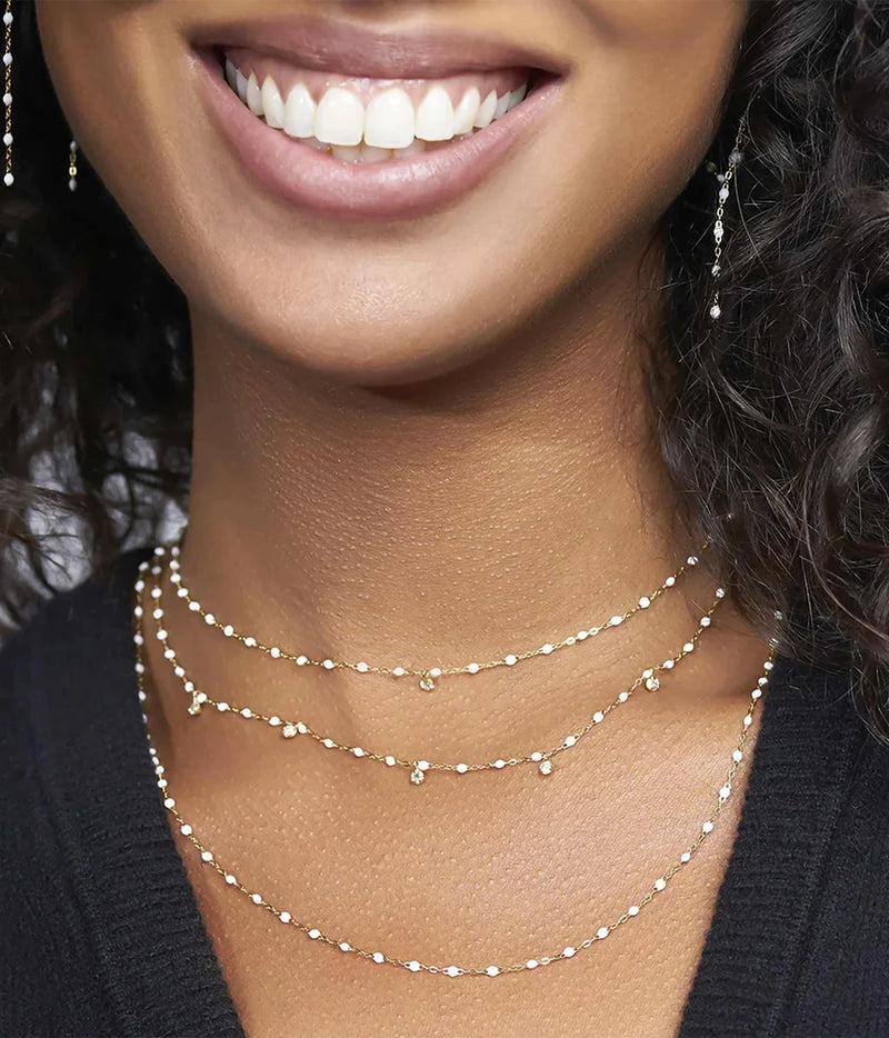 Close-up of woman's neck and smile wearing three layered white beaded gold necklaces with diamond charms, styled with matching drop earrings