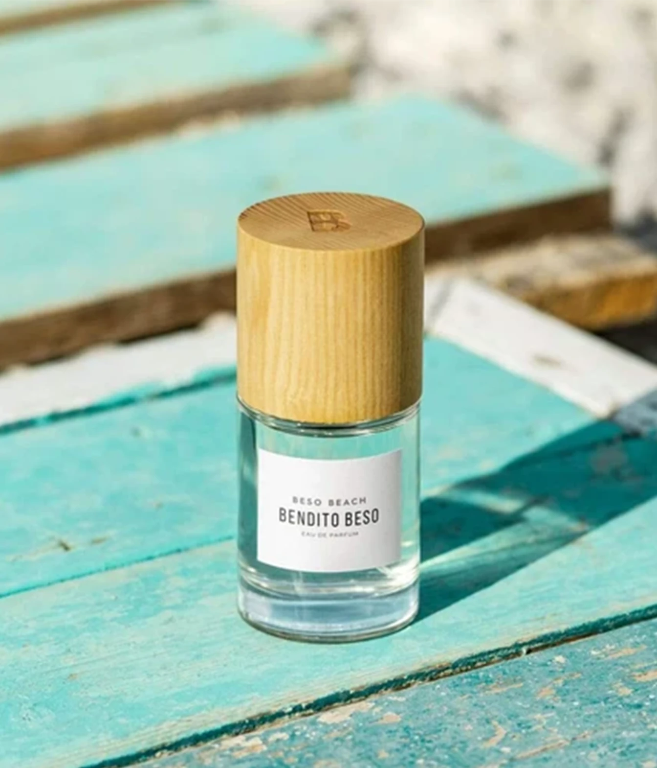 A clear glass perfume bottle with a light wood cap and a white label reading 'BESO BEACH BENDITO BESO', placed on a weathered turquoise wooden surface outdoors.