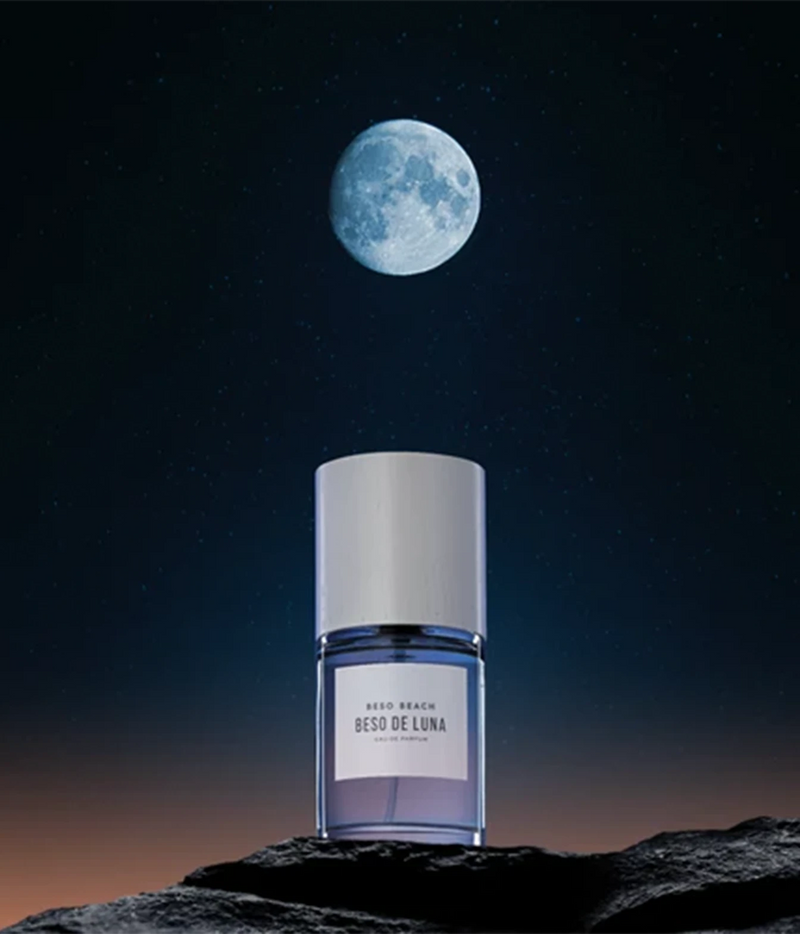 A bottle of Aedo Beach Besi de Luna body spray stands on a dark rocky surface against a night sky with a large moon and stars. The bottle is clear with a lavender-purple gradient liquid and a white cap.