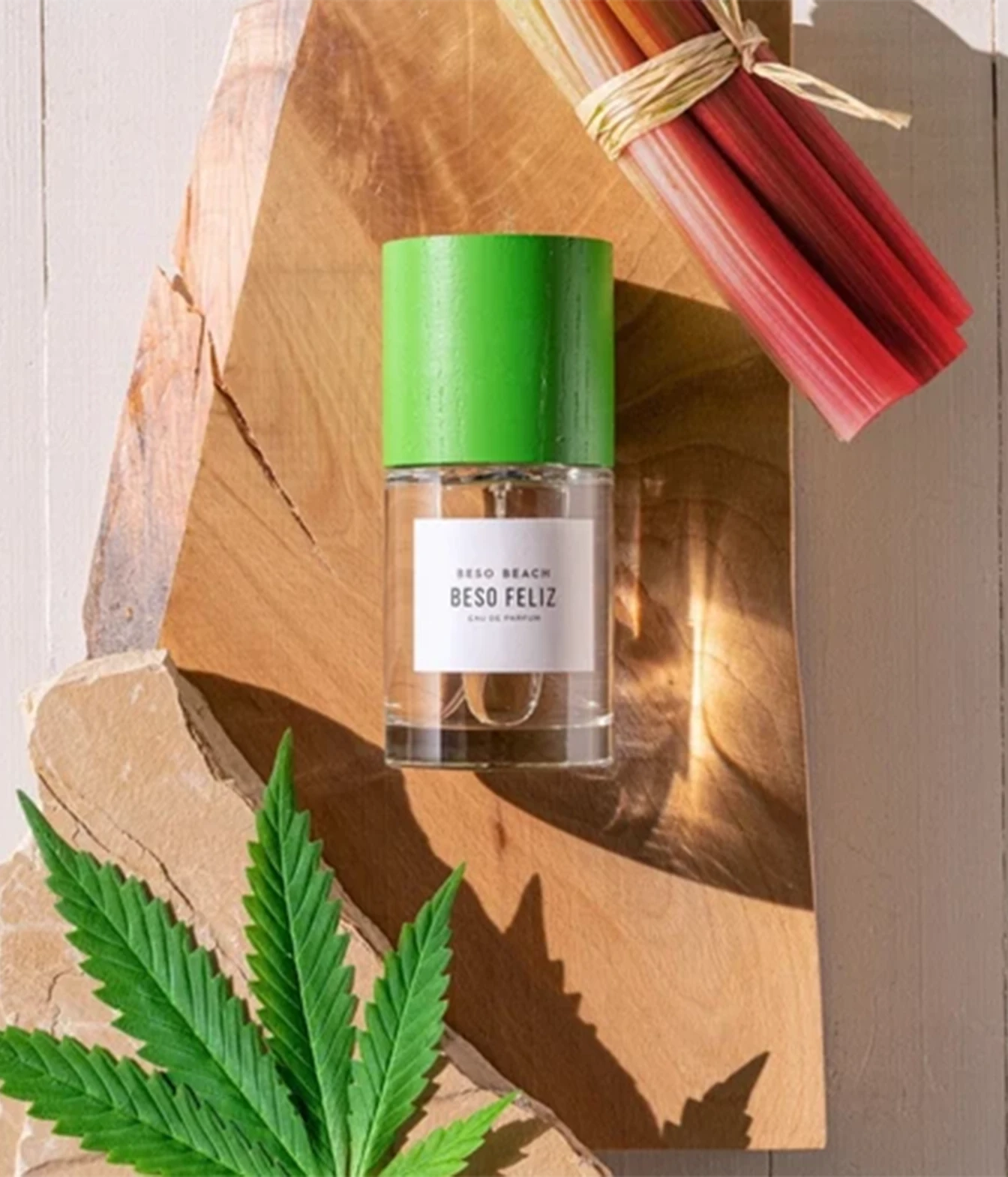 A clear glass bottle of Beso Beach Beso Feliz perfume with a bright green cap and white label, styled on a wooden board with a green leaf and red rhubarb in the background.
