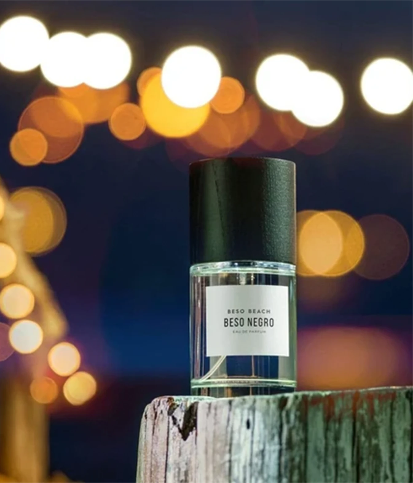 A bottle of Beso Negro Eau de Parfum with a black cap and white label, set on a weathered wooden block against a dark background with warm bokeh lights.