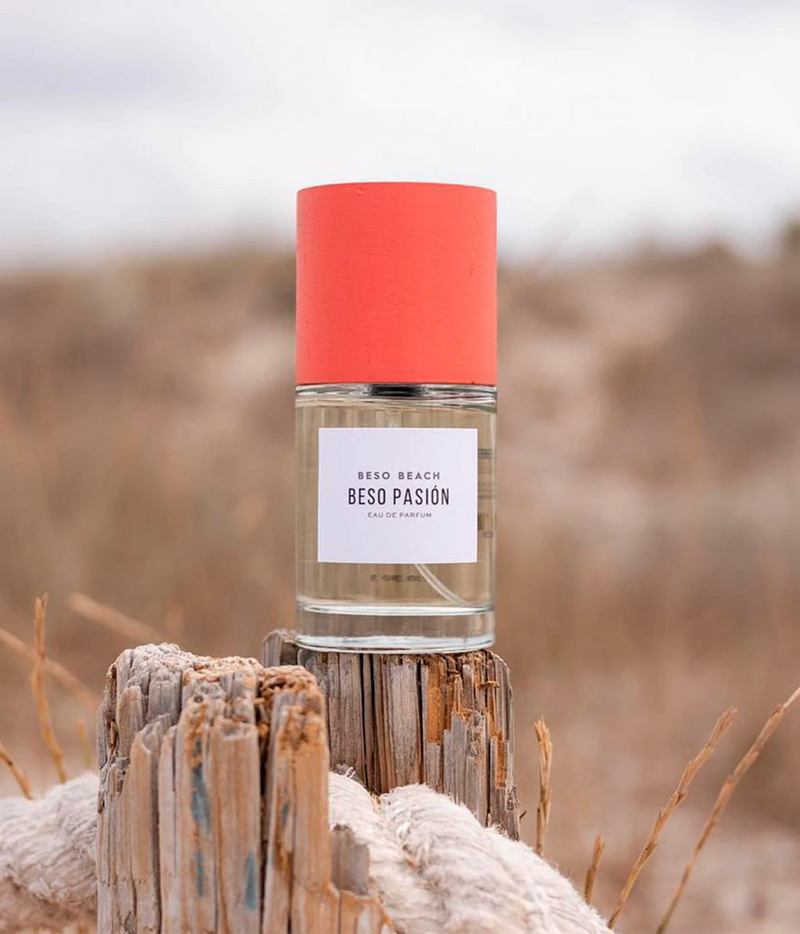 A bottle of Beso Beach Beso Pasion Eau de Parfum with a bright coral-red cap and a white label, set on a weathered wooden post with a blurred beach dune background.