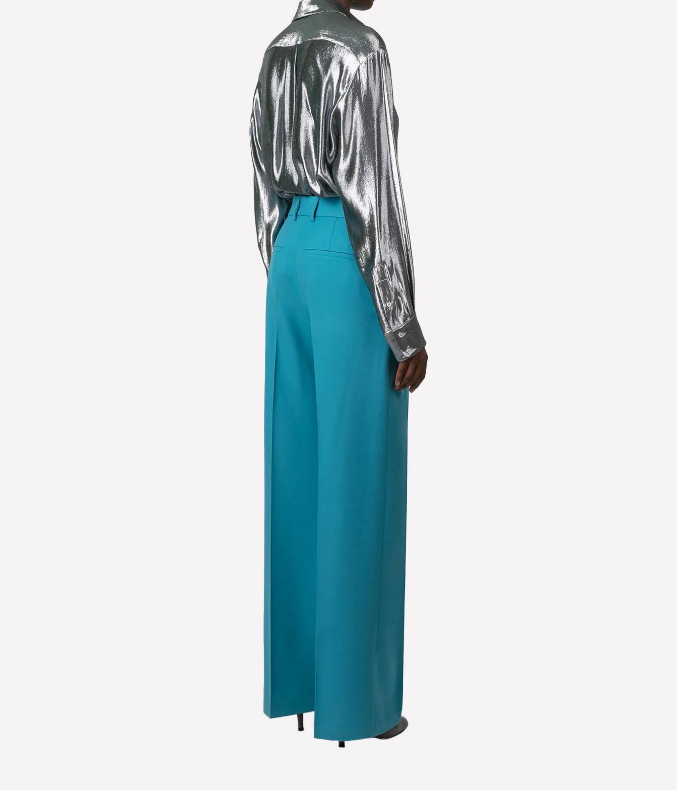 A model shown from the back wearing a shiny silver long-sleeve shirt and high-waisted, wide-leg turquoise blue trousers, set against a plain light gray background.