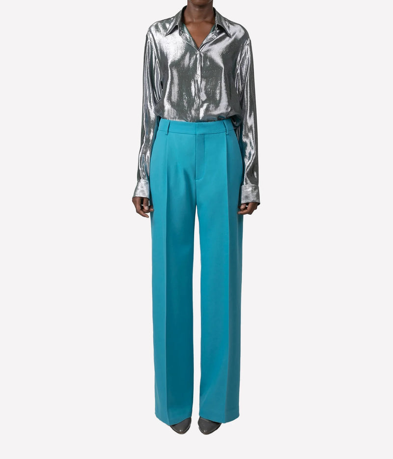 A model wearing a shiny silver satin button-down shirt and bright turquoise wide-leg trousers, shown from the neck down against a light gray background.