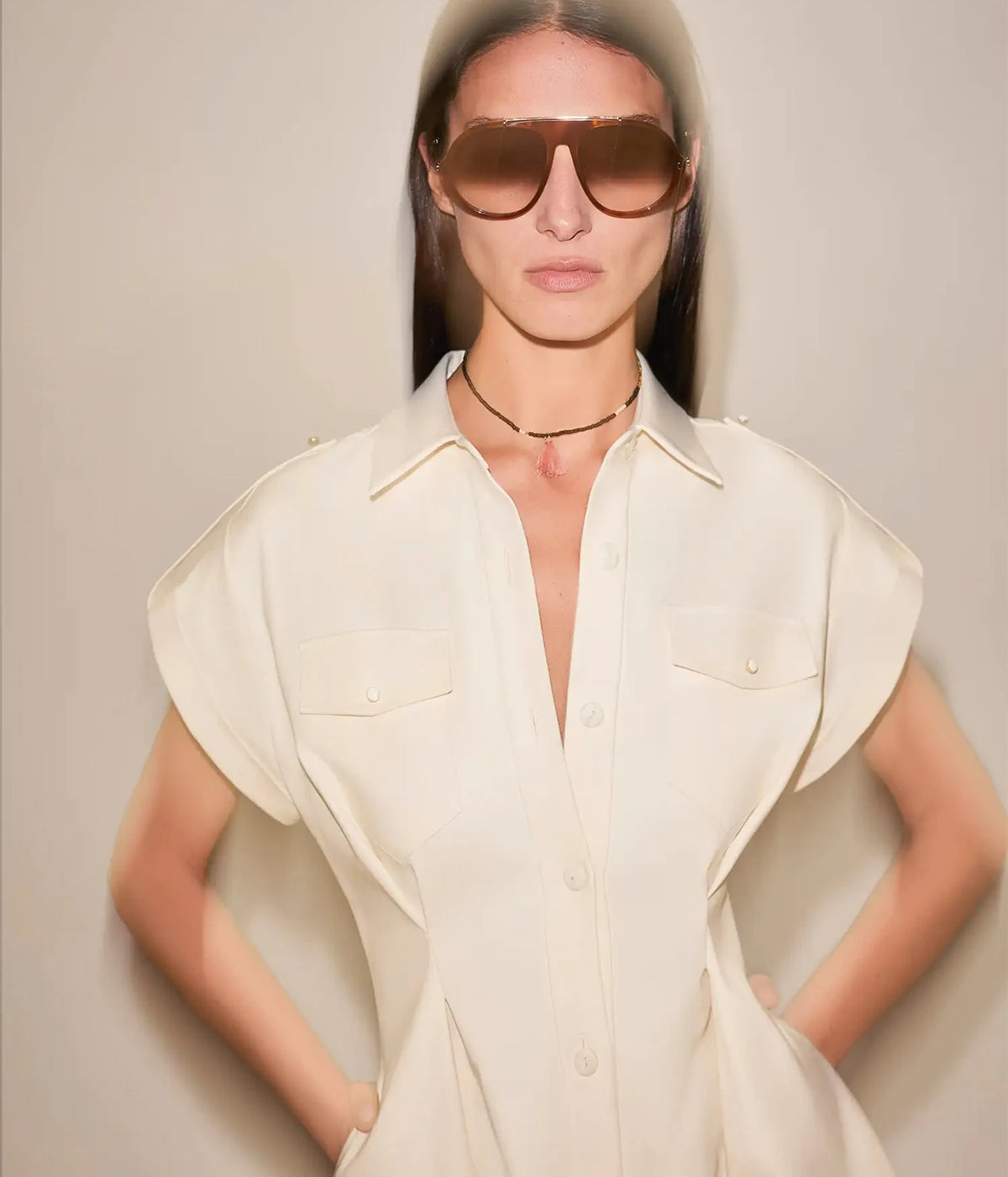 Close-up of a model wearing the Alexis Ceres Dress In Beige with hands on hips, showing the open collar, cap sleeves and fitted bodice, styled with brown sunglasses and a fine necklace