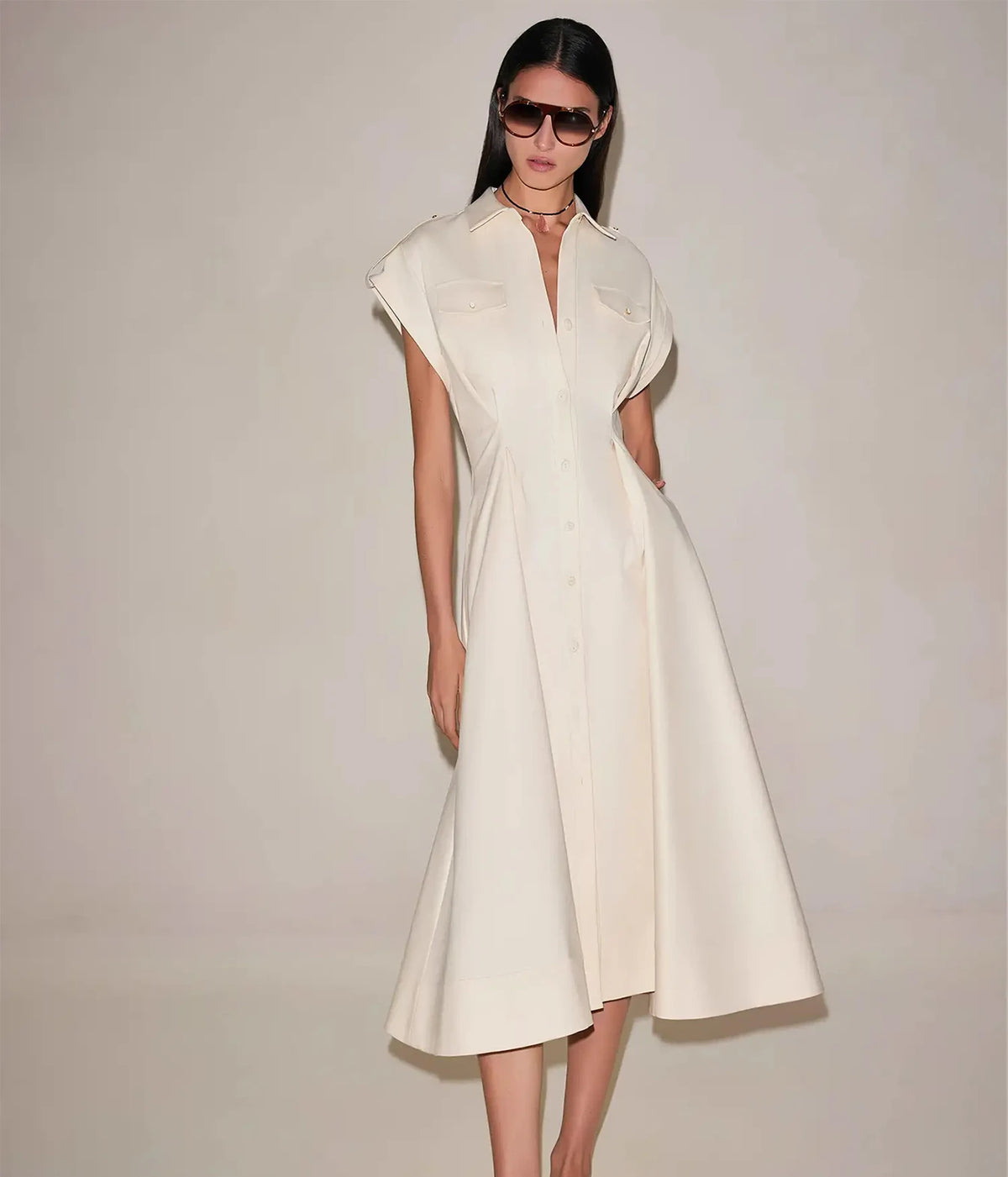 Full-length front view of a model wearing the Alexis Ceres Dress In Beige with brown sunglasses and a fine necklace, showing the open collar, cap sleeves, button front and full A-line midi skirt