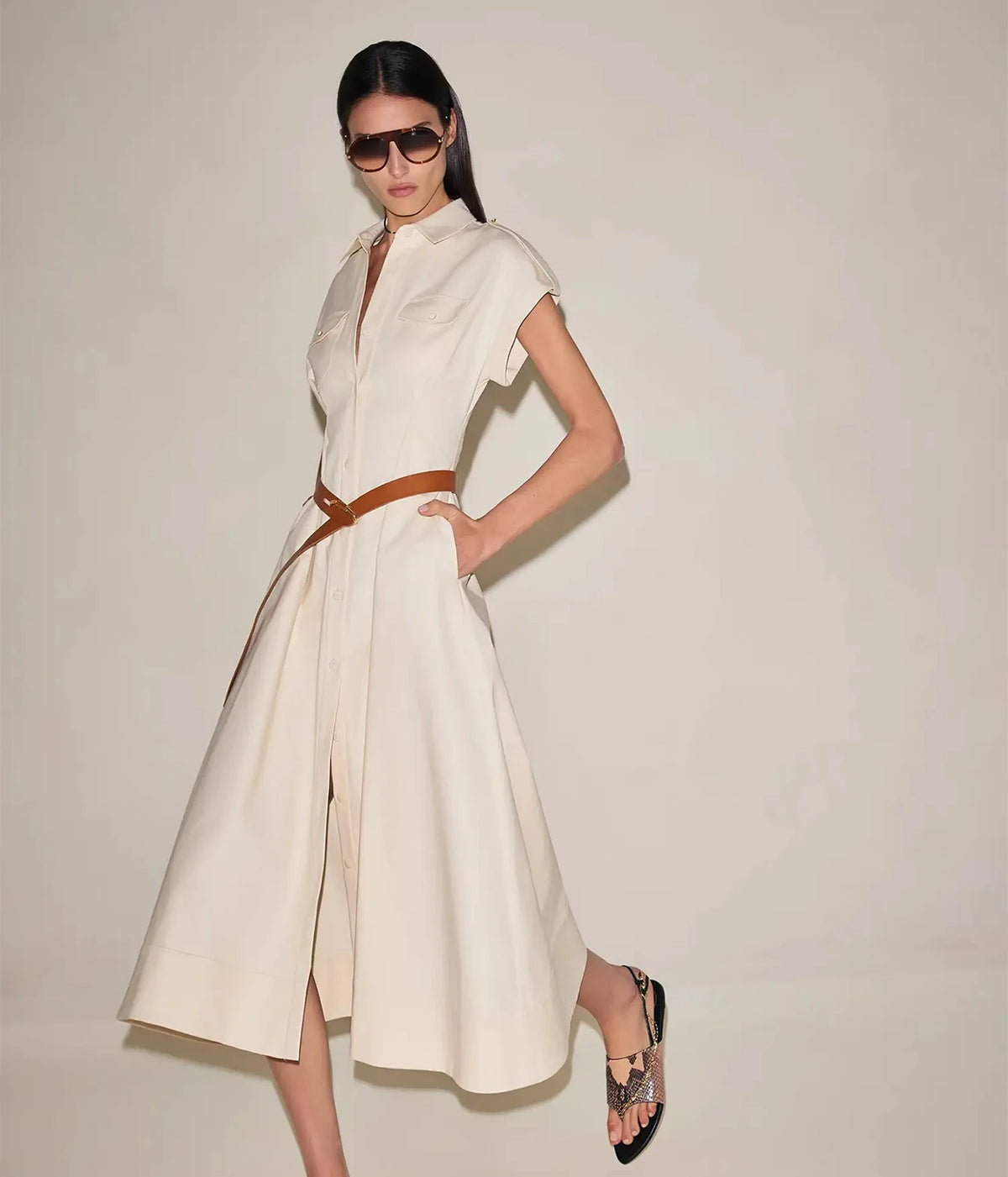 Model walking in the Alexis Ceres Dress In Beige cinched with a thin brown belt, styled with brown sunglasses and brown heeled sandals
