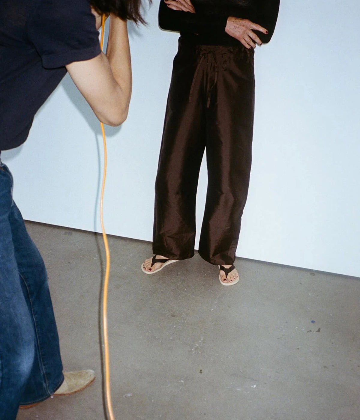 A person wearing dark chocolate brown wide-leg pants and black sandals stands against a plain light blue wall.