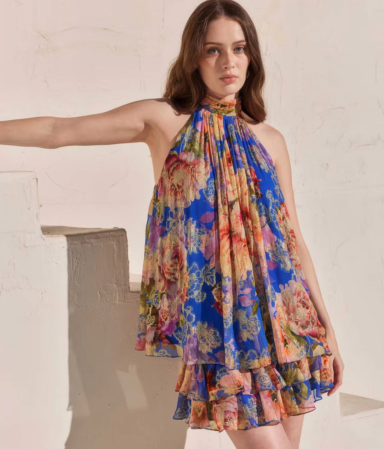 A woman models a sleeveless halter-neck mini dress with a vibrant multicolor floral print. The dress features a rich blue base with prominent pink, yellow, and orange floral motifs, and a tiered ruffled hem.