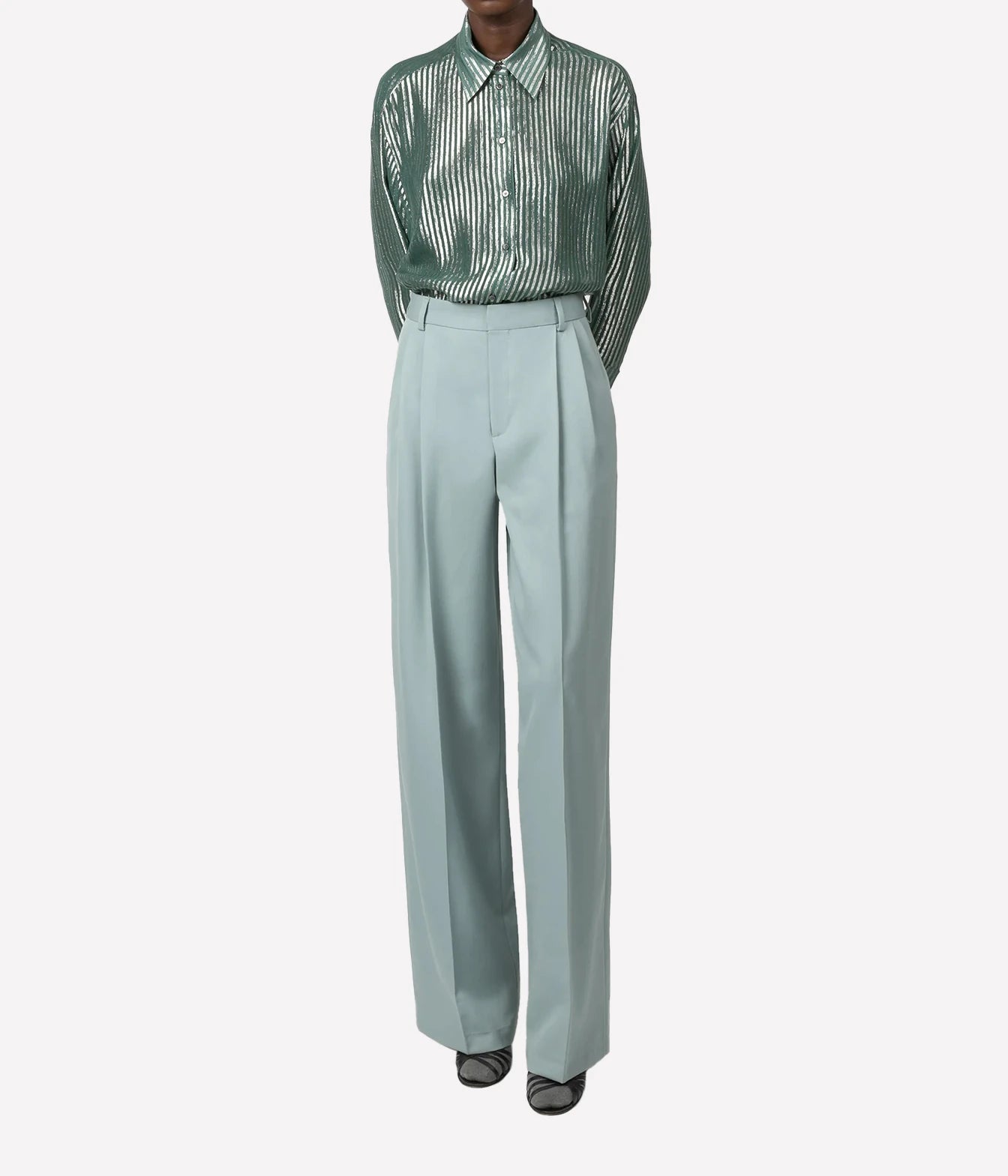A model is shown from the neck down wearing a green and white vertically striped long-sleeve button-up shirt tucked into high-waisted, wide-leg sage green trousers, paired with black pointed shoes, against a light gray background.