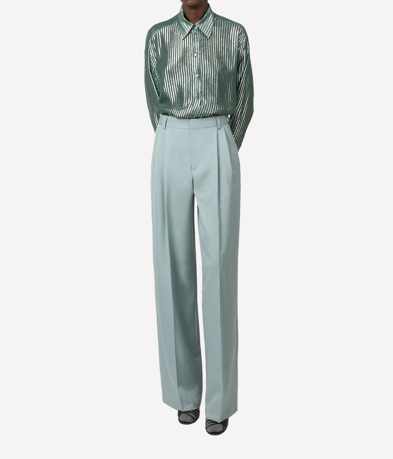 A model is shown from the neck down wearing a green and white vertically striped long-sleeve button-up shirt tucked into high-waisted, wide-leg sage green trousers, paired with black pointed shoes, against a light gray background.