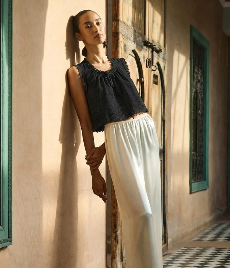 A woman leans against a sunlit beige wall beside a rustic wooden door, wearing a sleeveless black lace-trim top and flowing white wide-leg pants.
