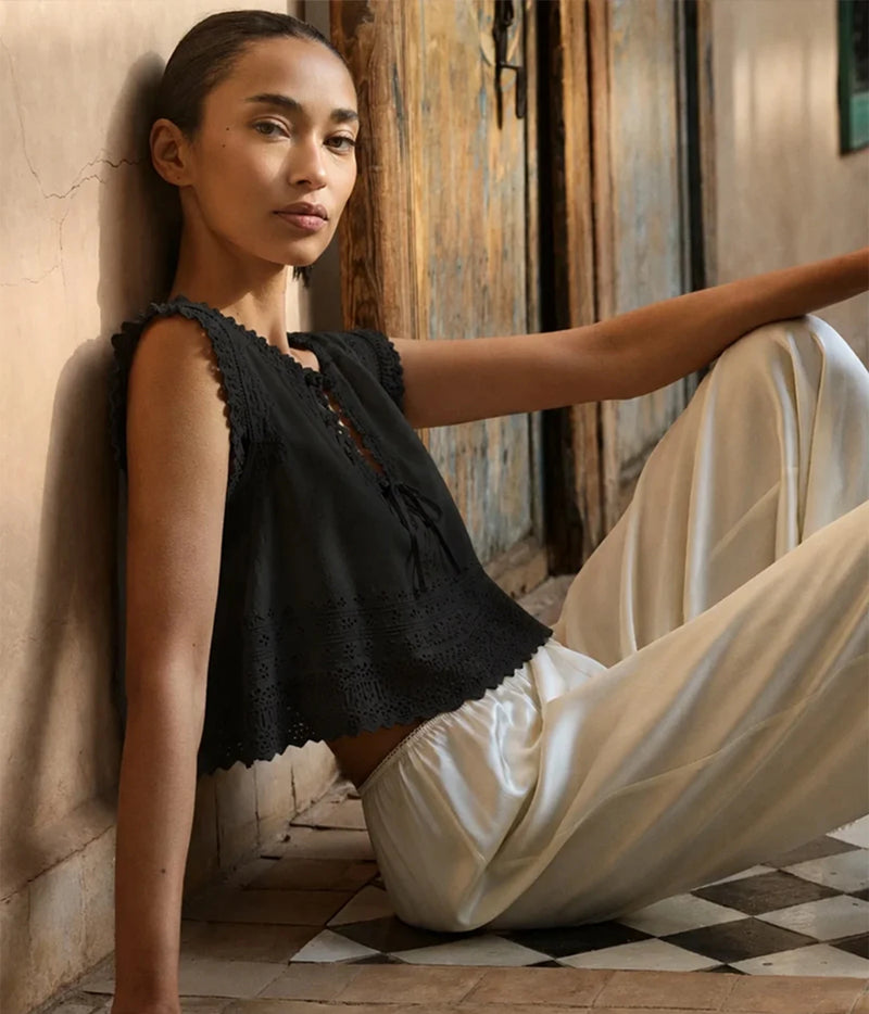 A woman sits on a checkered floor against a rustic wall, wearing a black sleeveless cropped top with lace trim and cream satin pants.