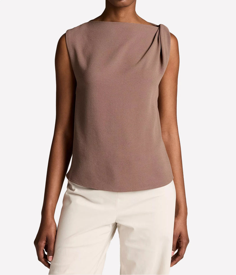 A woman wearing a sleeveless draped top in a muted taupe-brown color, paired with off-white pants, against a plain light gray studio background.