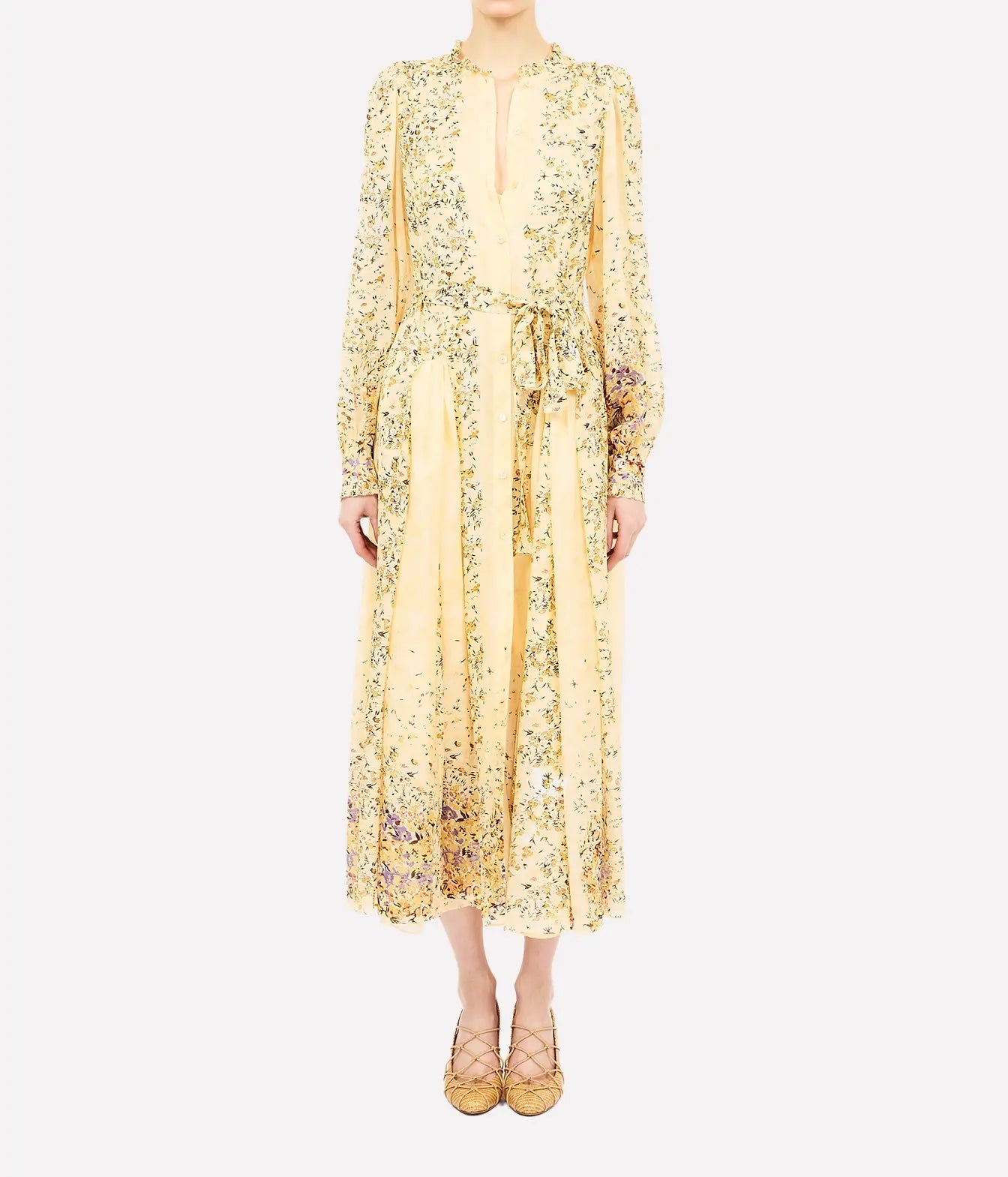 A model wearing a long-sleeved midi dress in pale yellow with a dense green and purple floral print, featuring a tie waist and a split neckline, paired with matching yellow lace-up flats.